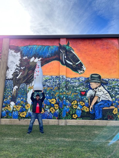 Blake Smith, Distribution Supervisor, hoists the Recognition Roadtrip Bottle in front of a cowboy themed mural in downtown Bryan that offers the advice, “Wherever you go, go with all your heart.”